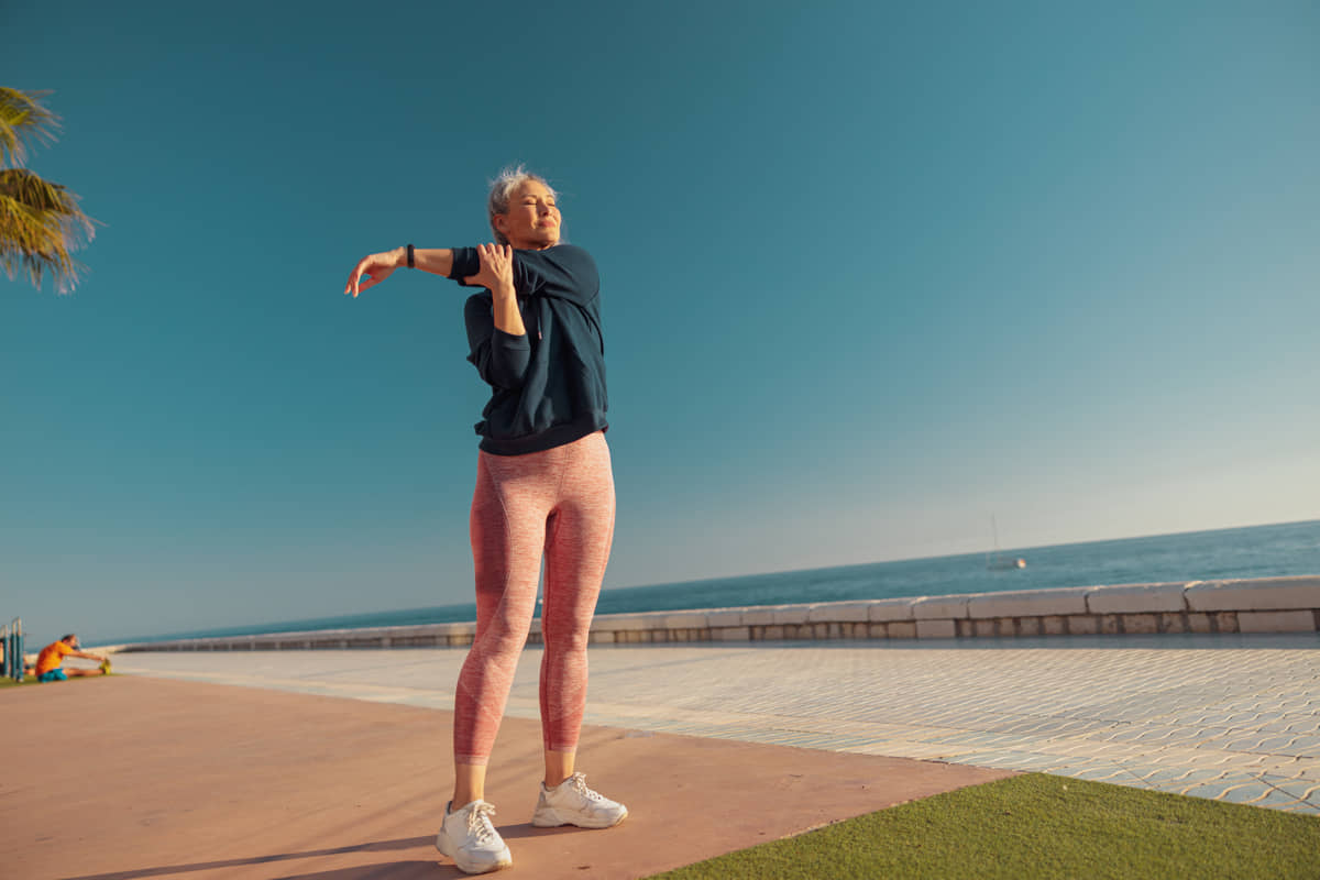 Silver-haired woman in a blue sweatshirt and pink leggings stretching her arm on a sunny seaside promenade.