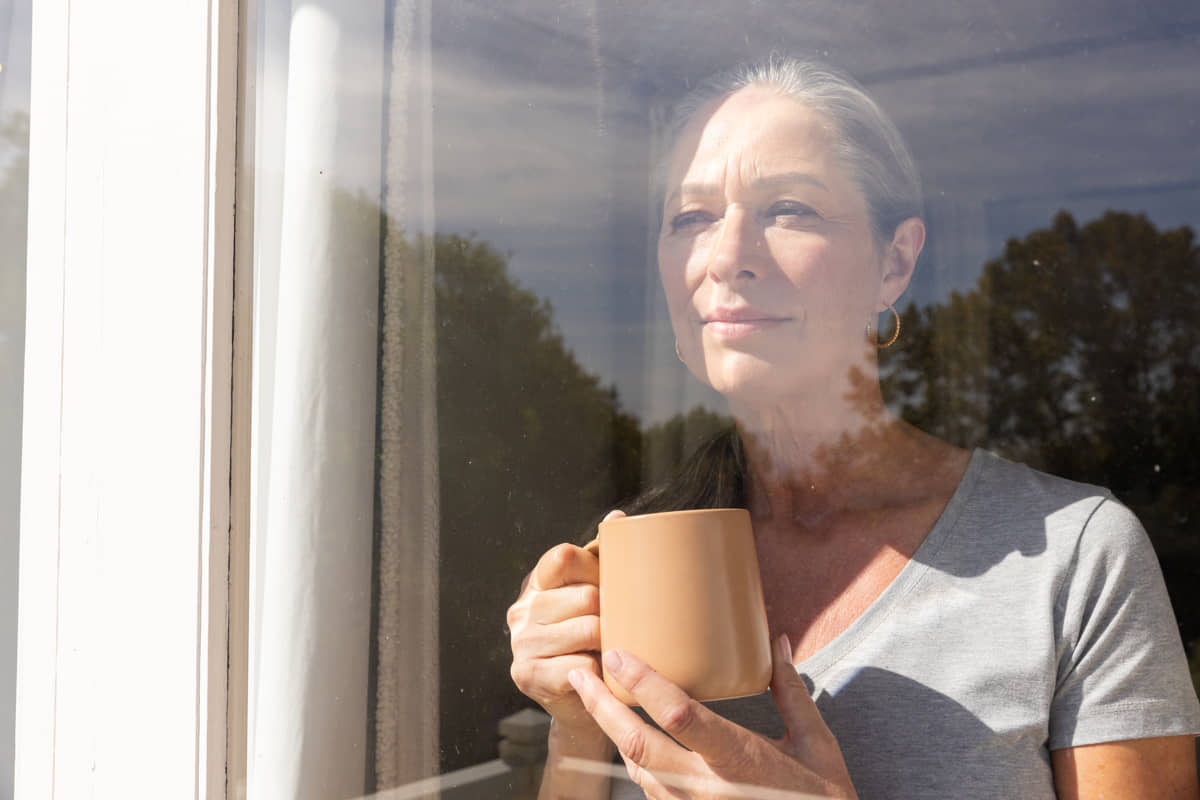 Silver-haired woman holding a mug and smiling as she looks out the window.