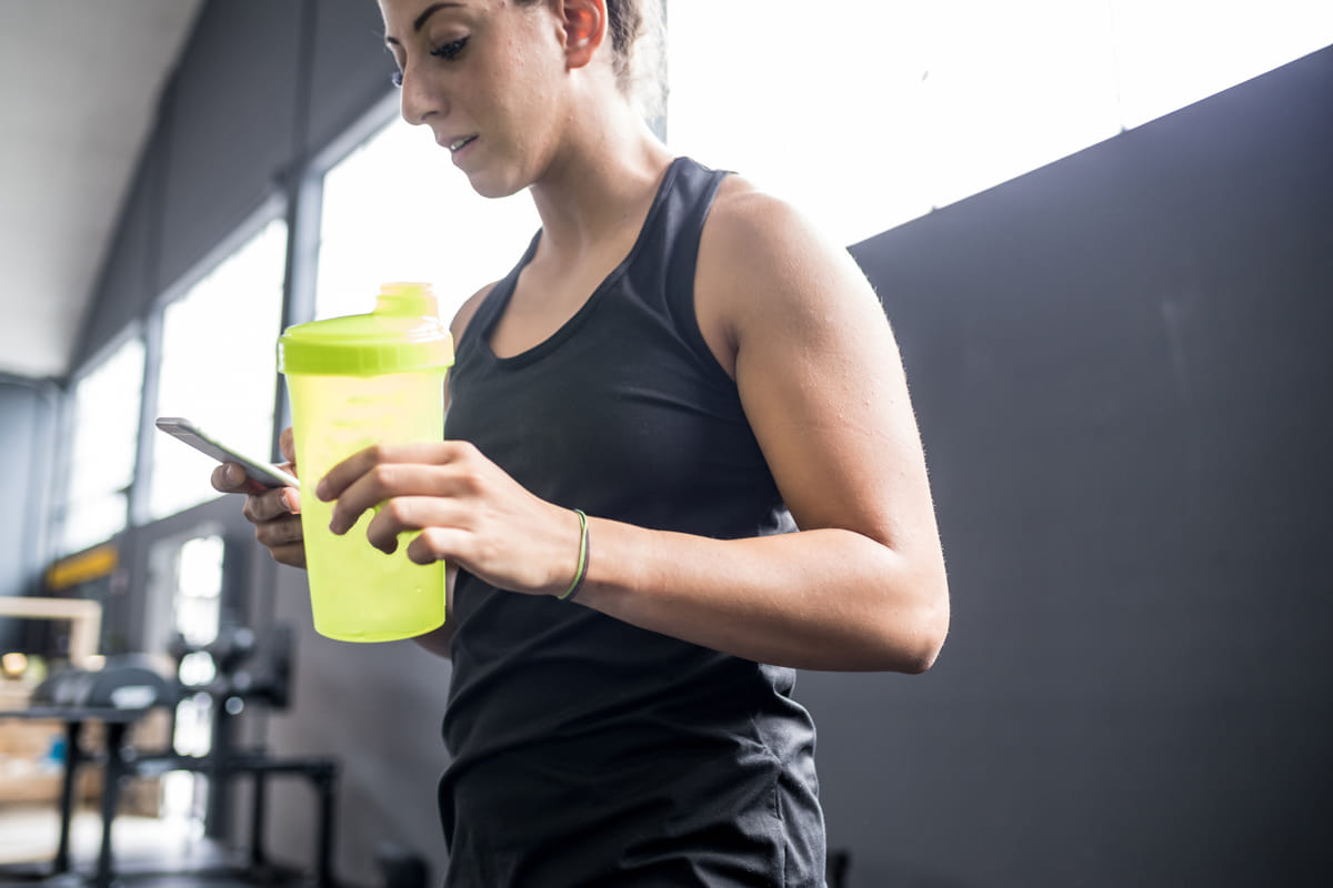 Woman in a gym drinking from a green shaker while checking a weight-loss tracking app on her phone.