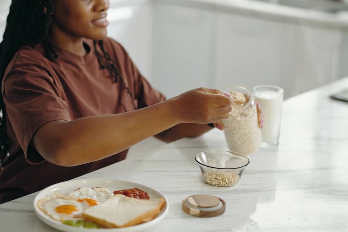 Woman in a brown t-shirt serving oats from a glass jar into a bowl, next to a breakfast plate of fried eggs, toast and a glass of milk.