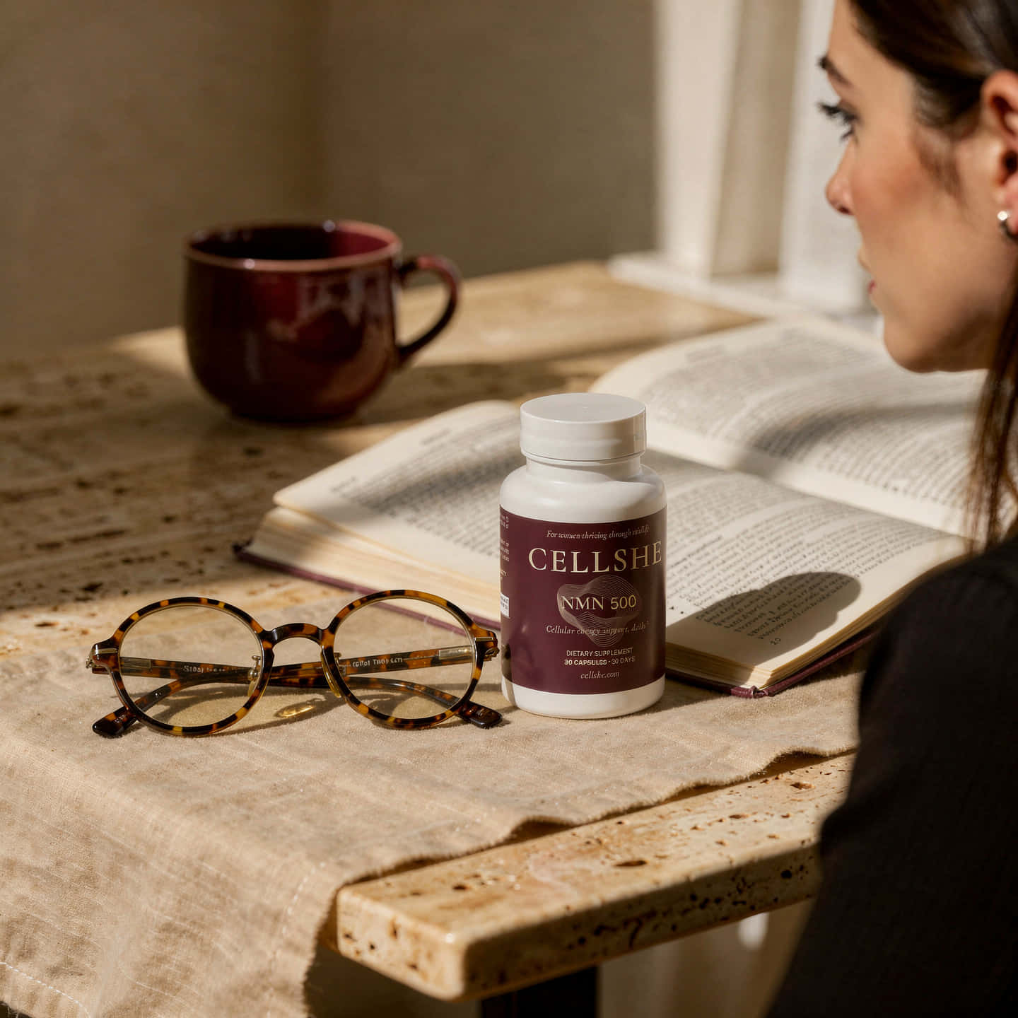 CELLSHE NMN 500 bottle on a table beside glasses, an open book, and a woman reading